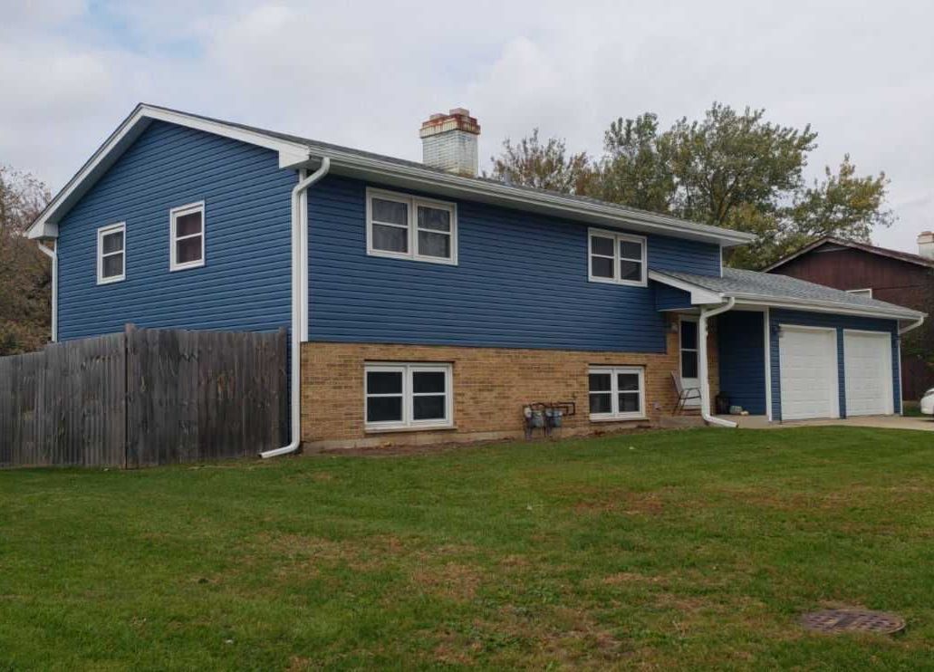 McHenry Avenue Crystal Lake IL after — blue vinyl siding, white trim, new roof, fascia, soffit, gutters, and new white garage doors